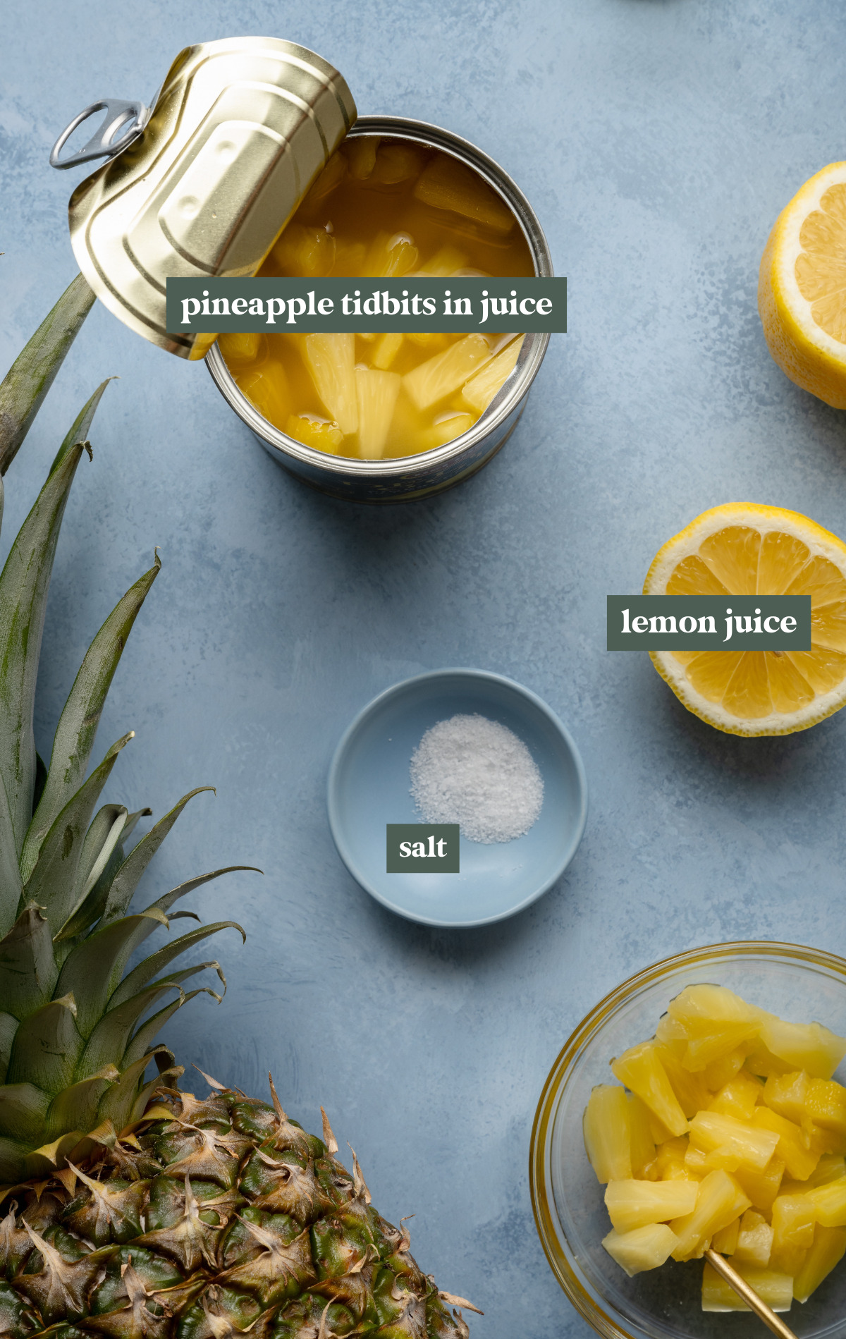 Flat lay of pineapple sorbet ingredients on a blue background, including a can of pineapple chunks in juice, lemon halves, a bowl of salt, a fresh pineapple, and a bowl of drained pineapple chunks.

