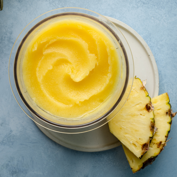 Pineapple sorbet in ninja creami pint container on white plate with two pineapple chunks garnishing the plate.