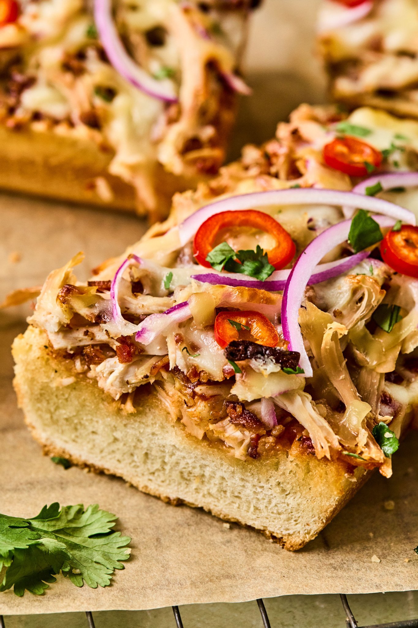 Close-up of a slice of BBQ chicken French bread pizza topped with melted cheese, barbecue sauce drizzle, red onion, and sliced red chili peppers.