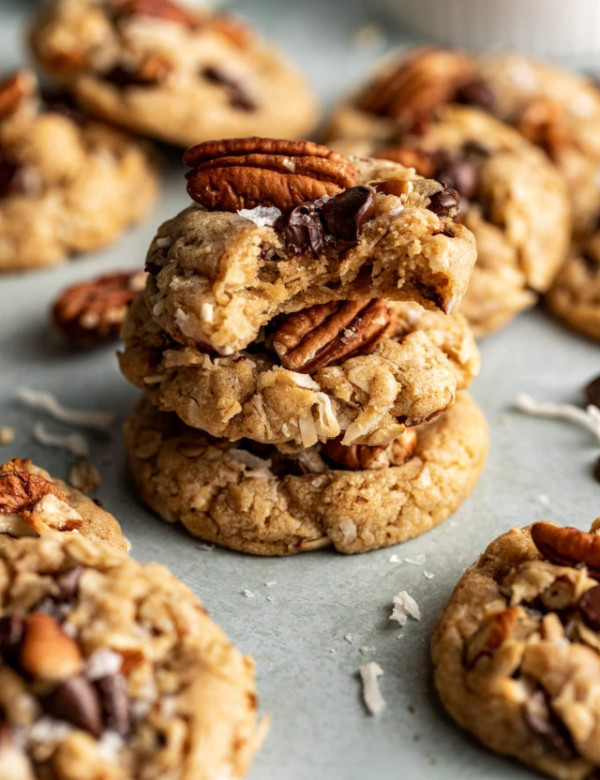 Three cowboy cookies stacked on top of each other on plate with the top cookie having a bite taken out of the top and more cookies scattered around the stack.