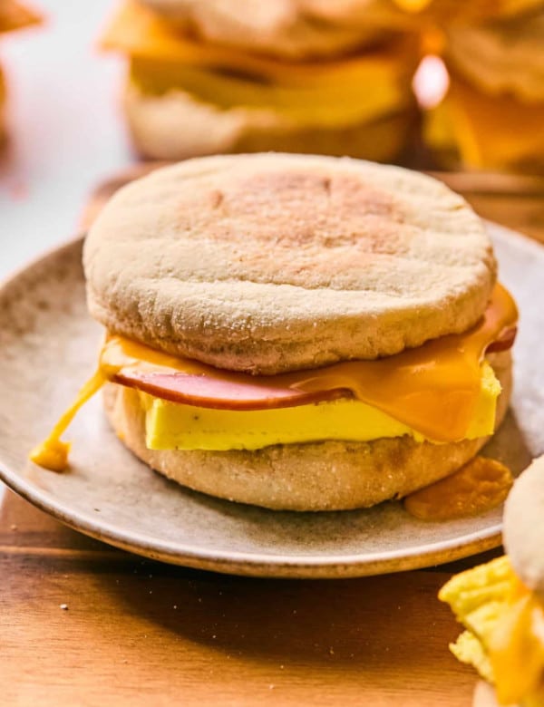 A breakfast sandwich on a ceramic plate made with an English muffin, cheddar cheese, folded egg, and a round slice of ham. The cheese is melted and slightly dripping from the edge.