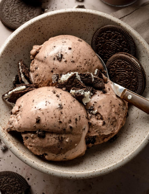 A speckled ceramic bowl filled with three scoops of creamy chocolate Oreo ice cream, topped with crushed Oreo cookie pieces. Whole Oreo cookies are placed on the side and in the background, with a metal spoon resting in the bowl. The ice cream is smooth with visible cookie bits throughout.