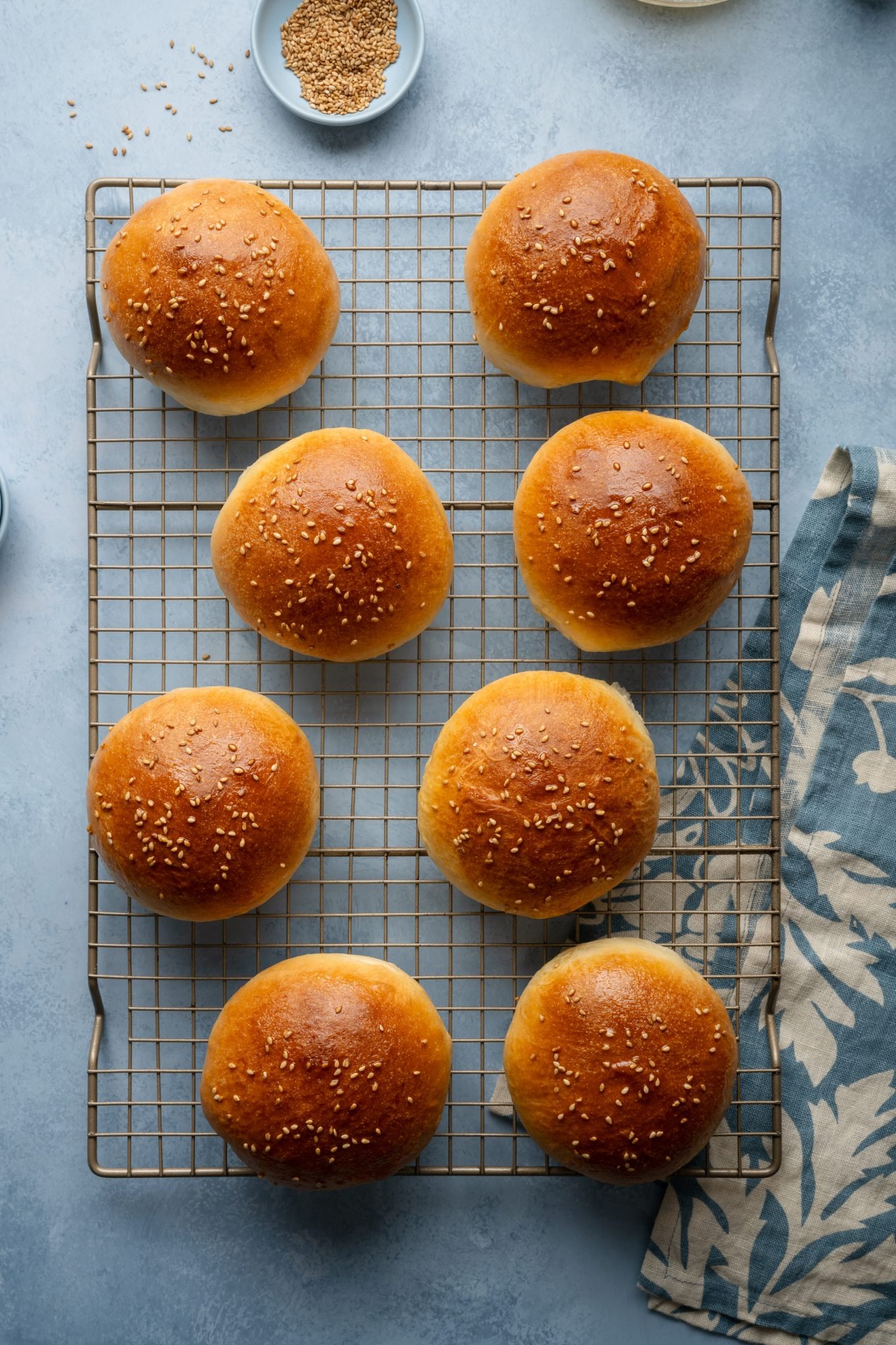 Freshly baked sesame seed hamburger buns cooling on a wire rack, with a blue floral kitchen towel and a small dish of sesame seeds nearby.