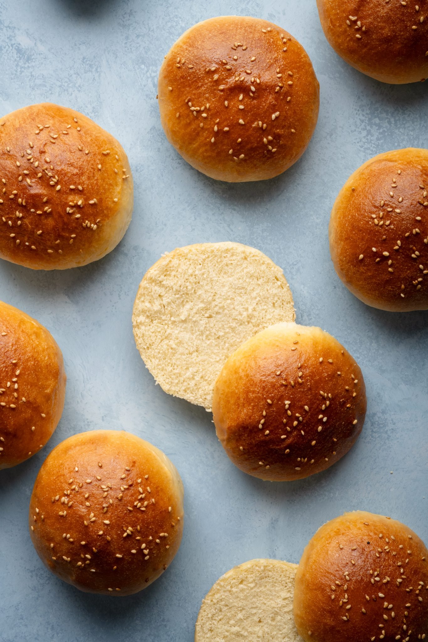 Top-down view of several golden hamburger buns with one sliced open to reveal its soft, fluffy interior.