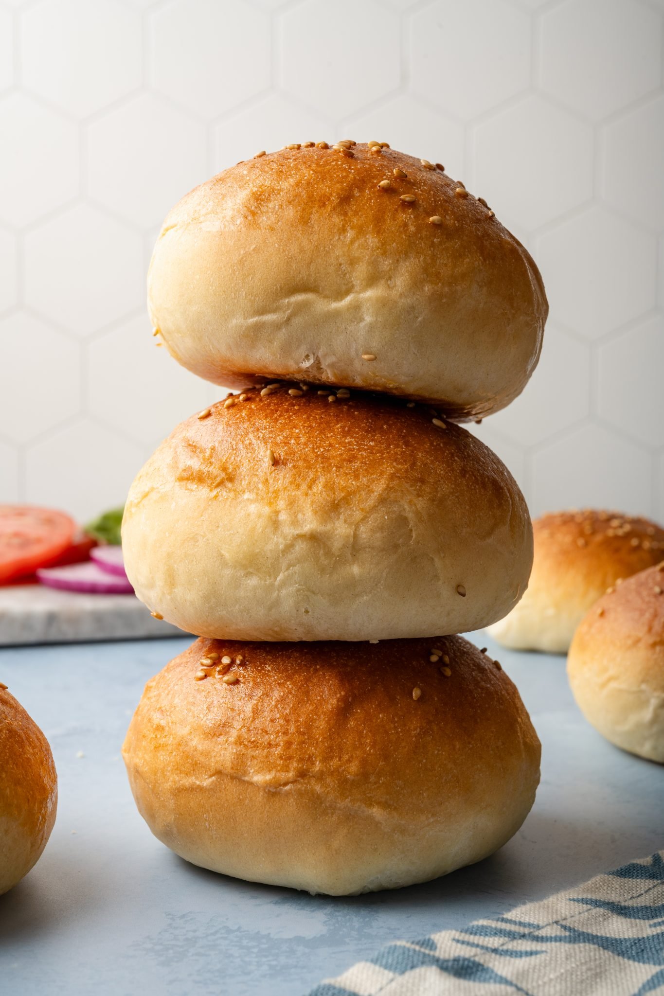 Three hamburger buns stacked tall on top of each other, showing their golden crust and pillowy sides with sesame seeds scattered on top.
