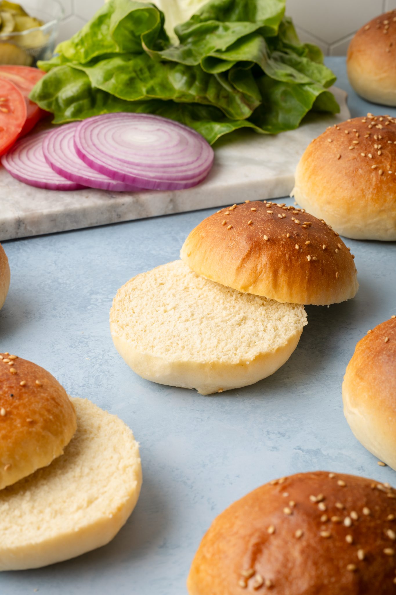 Open-faced hamburger buns surrounded by sliced tomatoes, red onion, and leafy lettuce on a marble board in the background.