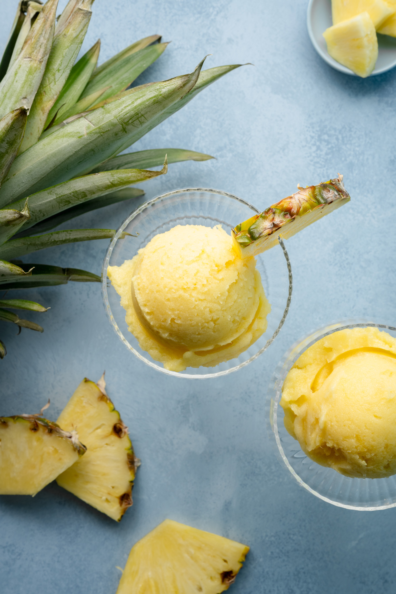 Overhead view of two scoops of creamy pineapple sorbet in glass dessert cups, each garnished with a fresh pineapple wedge, surrounded by pineapple leaves and slices.