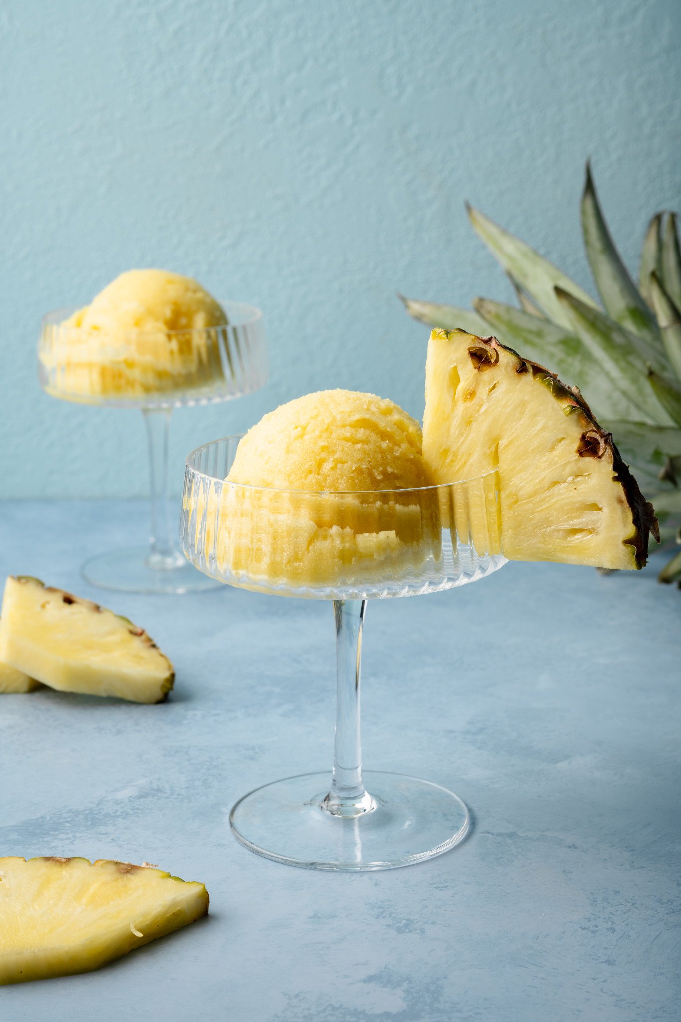 Close-up of pineapple sorbet served in a clear coupe glass with a thick pineapple wedge on the rim, placed on a blue background with a pineapple crown nearby.