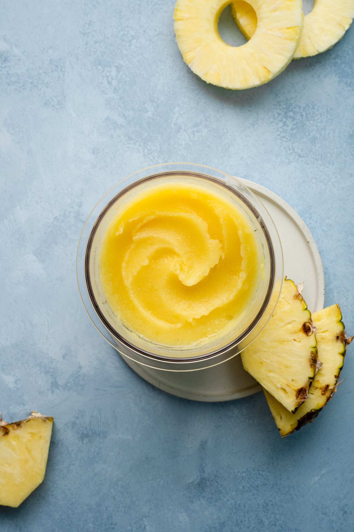 Top-down view of swirled pineapple sorbet in a Ninja Creami pint container, placed on a small plate beside pineapple slices and leaves.