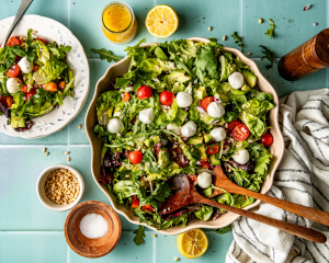 Favorite everyday salad tossed in large bowl with wooden spoons and plate of salad beside it. Jar of dressing and extra pine nuts and salt surround the salad.