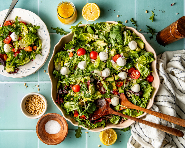 Favorite everyday salad tossed in large bowl with wooden spoons and plate of salad beside it. Jar of dressing and extra pine nuts and salt surround the salad.