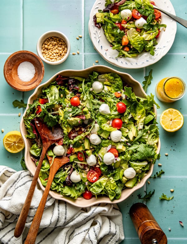 Large salad bowl with every day salad tossed together with two wooden spoons and one serving of salad plated on small plate with fork on table. There is extra salad dressing, pine nuts and salt on table as well.