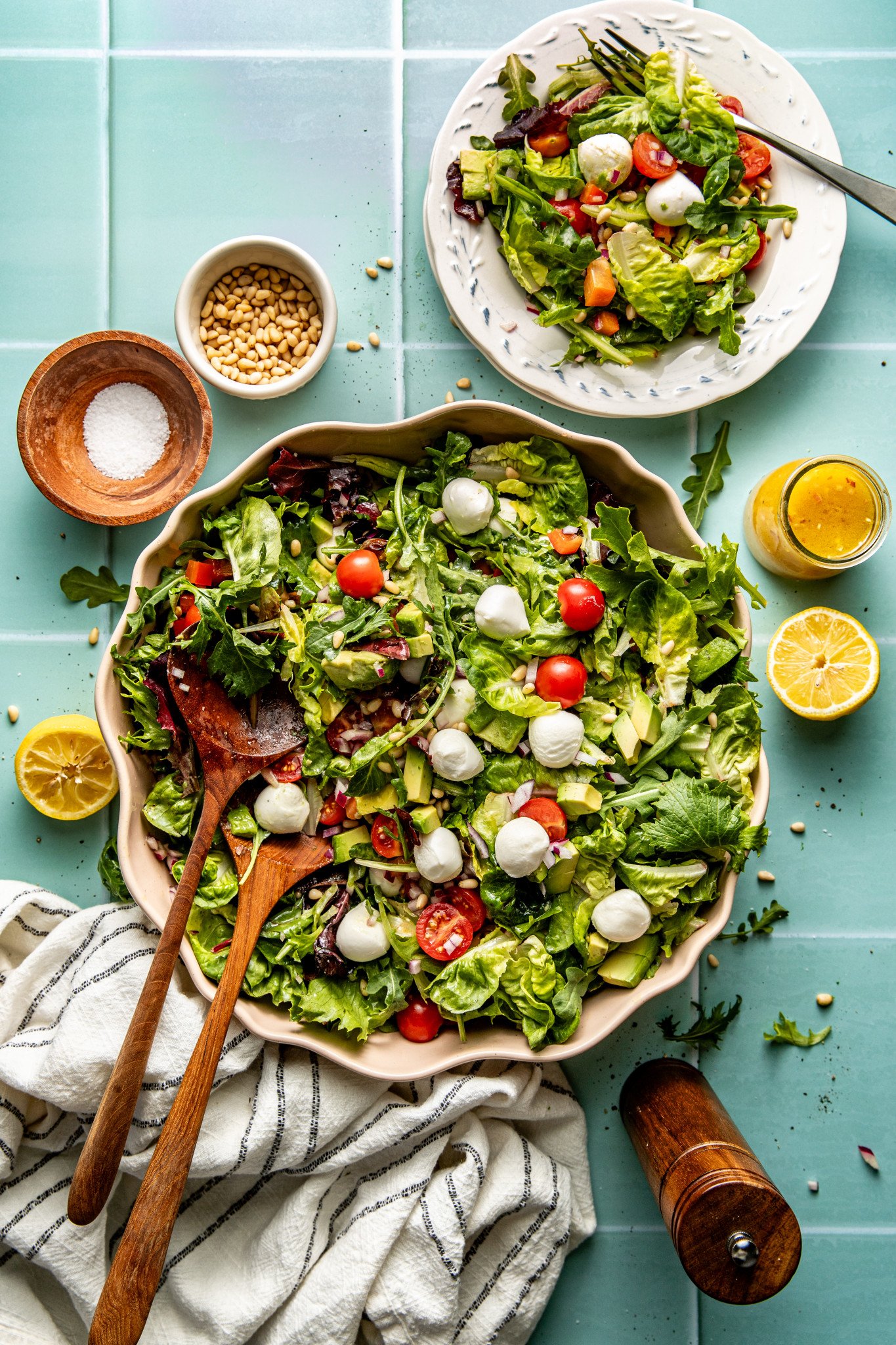 Large salad bowl with every day salad tossed together with two wooden spoons and one serving of salad plated on small plate with fork on table. There is extra salad dressing, pine nuts and salt on table as well.