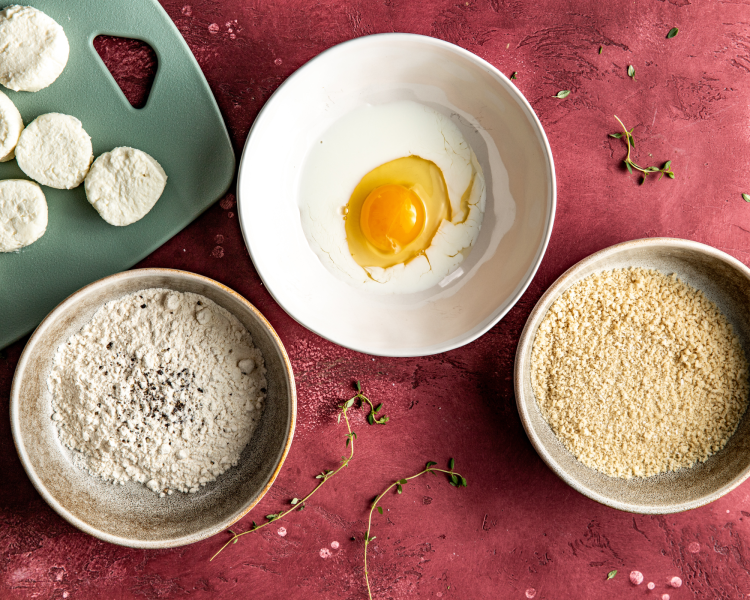 Three bowls set up for dredging station, one with flour, one with egg and milk, and one with panko bread crumbs for fried goat cheese.