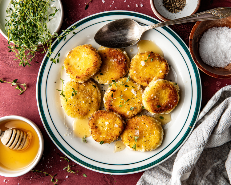 Eight goat cheese medallions on plate after being fried drizzled with honey and spoon on side of plate.