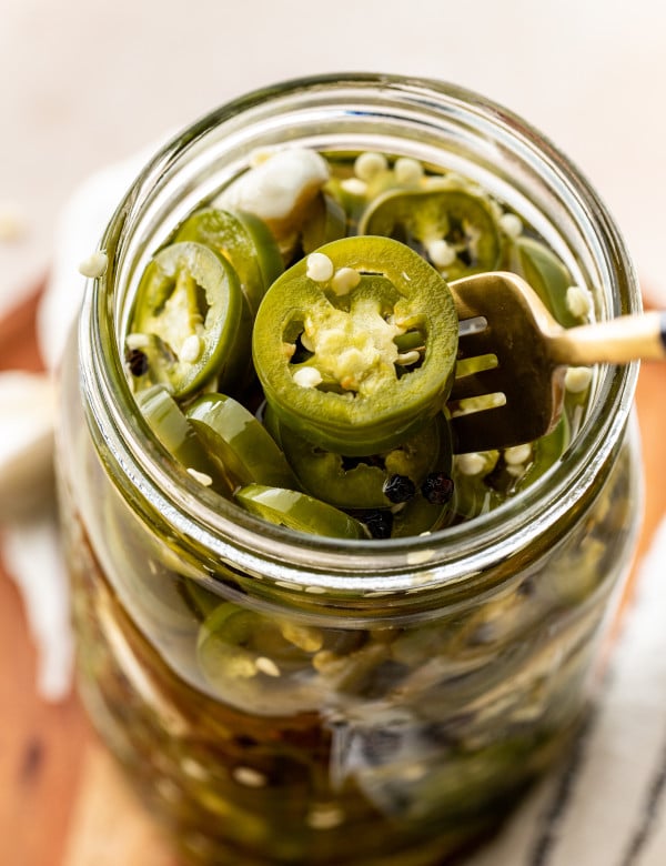 Pickled jalapenos in glass mason jar with gold fork lifting jalapeno out of jar.
