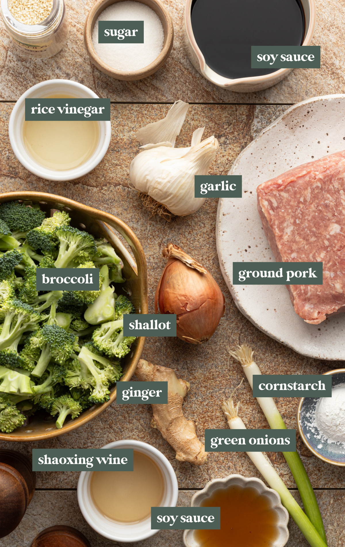 Overhead view of raw stir fry ingredients neatly arranged on a stone counter, including ground pork, broccoli florets, garlic, shallot, ginger, green onion, red bell pepper, soy sauce, vinegar, sugar, sesame seeds, and cornstarch.