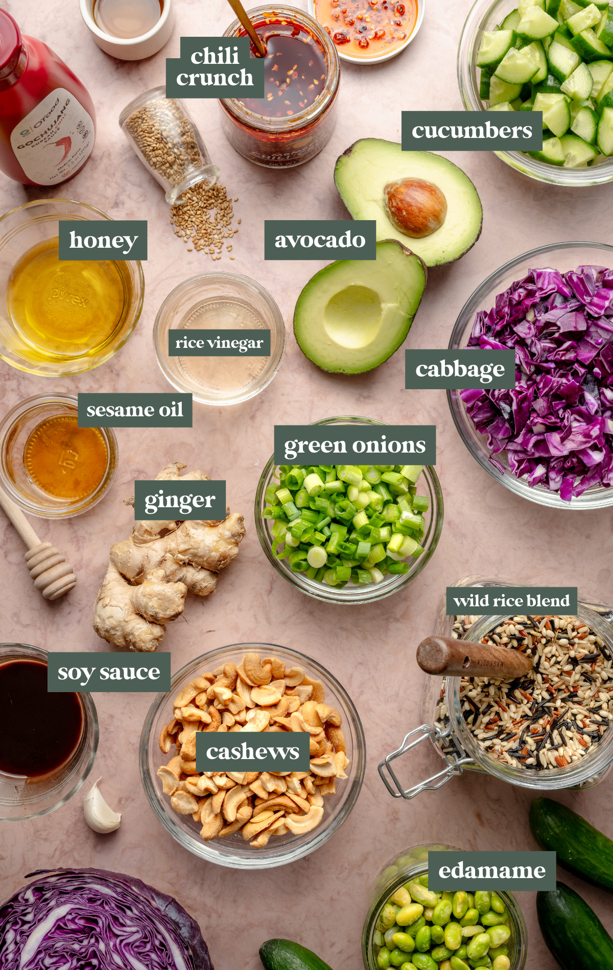 Flat lay of fresh crispy rice salad ingredients on a pink surface, including wild rice, chopped cucumbers, avocado halves, purple cabbage, edamame, green onions, cashews, ginger, garlic, sesame seeds, and sauces in small bowls.

