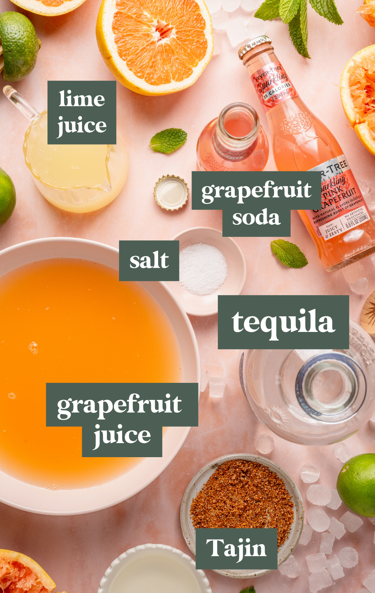 Overhead shot of Paloma cocktail ingredients including halved grapefruits, limes, grapefruit soda, lime juice, chili-lime salt, a bowl of orange-hued juice, and fresh mint on a pink surface with scattered ice.