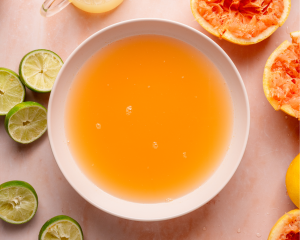 Overhead shot of a large bowl filled with fresh grapefruit-lime juice. Squeezed citrus halves, lime slices, and a small pitcher of juice surround the bowl on a pink marble surface.