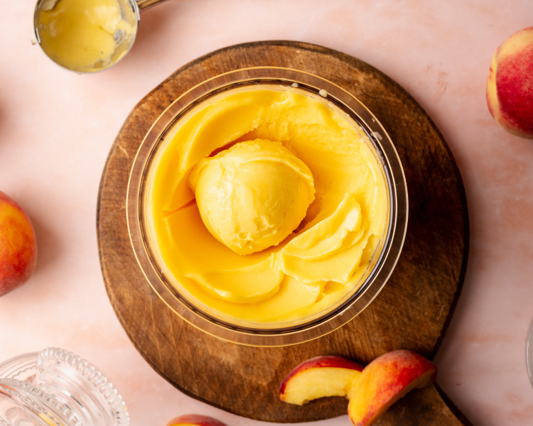Peach sorbet in ninja creami pint container scooped in container on top of wood cutting board with peaches on the side of the board.