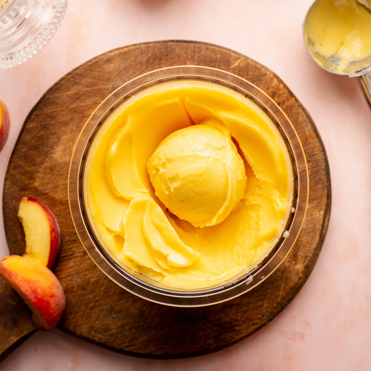 Peach sorbet in ninja creami pint container scooped in container on top of wood cutting board with peaches on the side of the board.