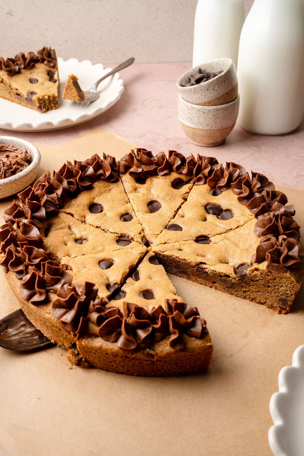 A decorated chocolate chip cookie cake with piped chocolate frosting is partially sliced, with one slice served on a plate in the background next to bottles of milk and stacked ceramic bowls.