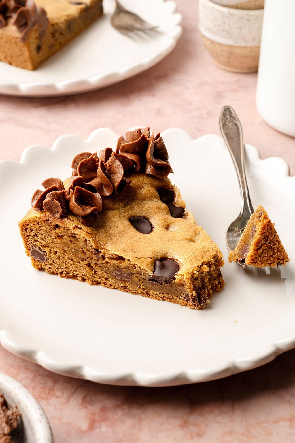 A slice of chocolate chip cookie cake on a white plate with chocolate frosting along the edge and a small piece on a fork ready to eat.