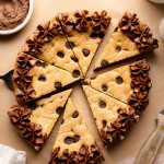 A sliced chocolate chip cookie cake decorated with piped chocolate frosting along the edges, with a cake server lifting one slice slightly away from the whole.