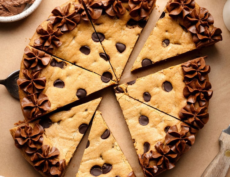 A sliced chocolate chip cookie cake decorated with piped chocolate frosting along the edges, with a cake server lifting one slice slightly away from the whole.