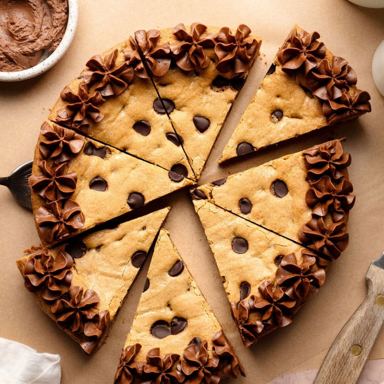 A sliced chocolate chip cookie cake decorated with piped chocolate frosting along the edges, with a cake server lifting one slice slightly away from the whole.
