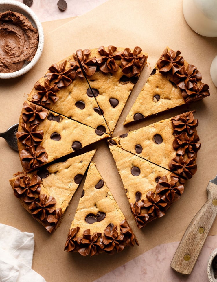 A sliced chocolate chip cookie cake decorated with piped chocolate frosting along the edges, with a cake server lifting one slice slightly away from the whole.