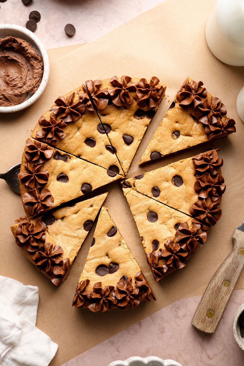 A sliced chocolate chip cookie cake decorated with piped chocolate frosting along the edges, with a cake server lifting one slice slightly away from the whole.