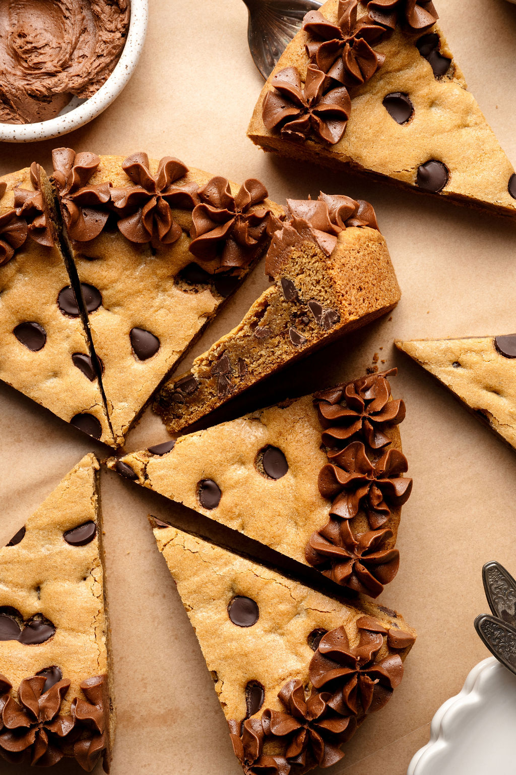 A close-up of a chocolate chip cookie cake with a slice pulled out, revealing a dense, chocolate-studded interior and piped chocolate frosting along the edge.