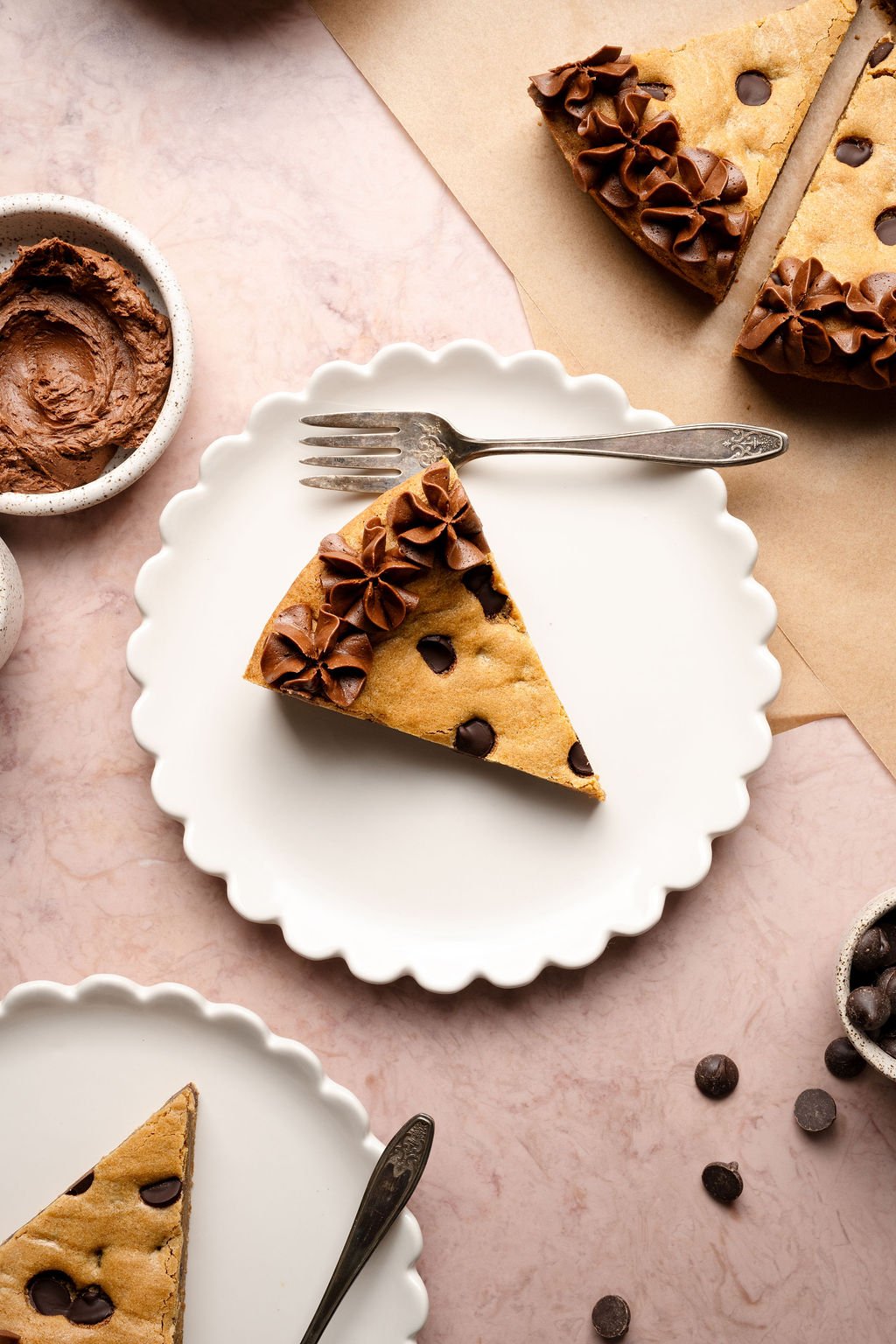 A single slice of chocolate chip cookie cake with chocolate frosting served on a white scalloped plate with a vintage fork, surrounded by bowls of frosting and chocolate chips.