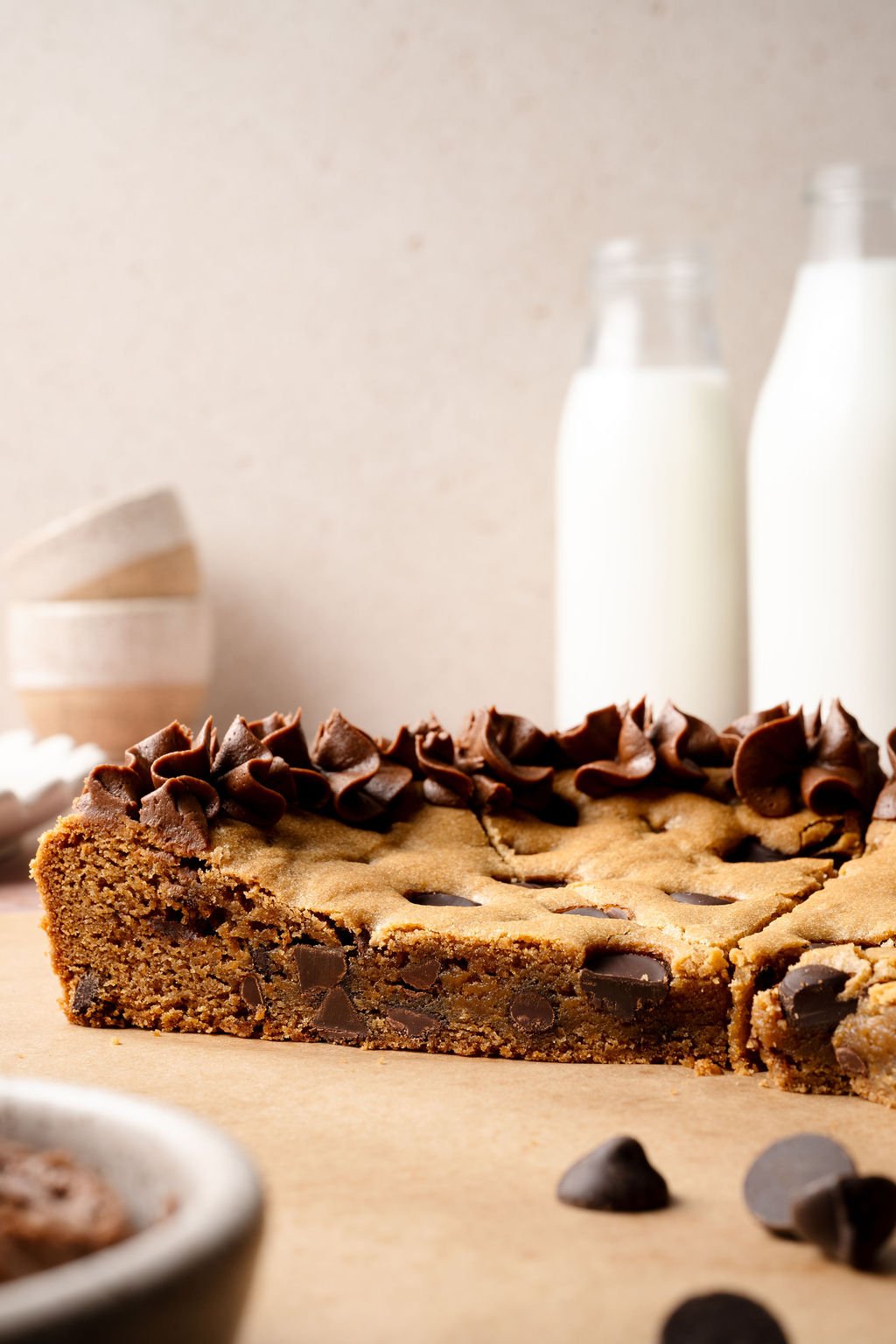 A side view of the chocolate chip cookie cake showing the thick, chewy interior filled with chocolate chips, topped with decorative chocolate frosting and two bottles of milk in the background.