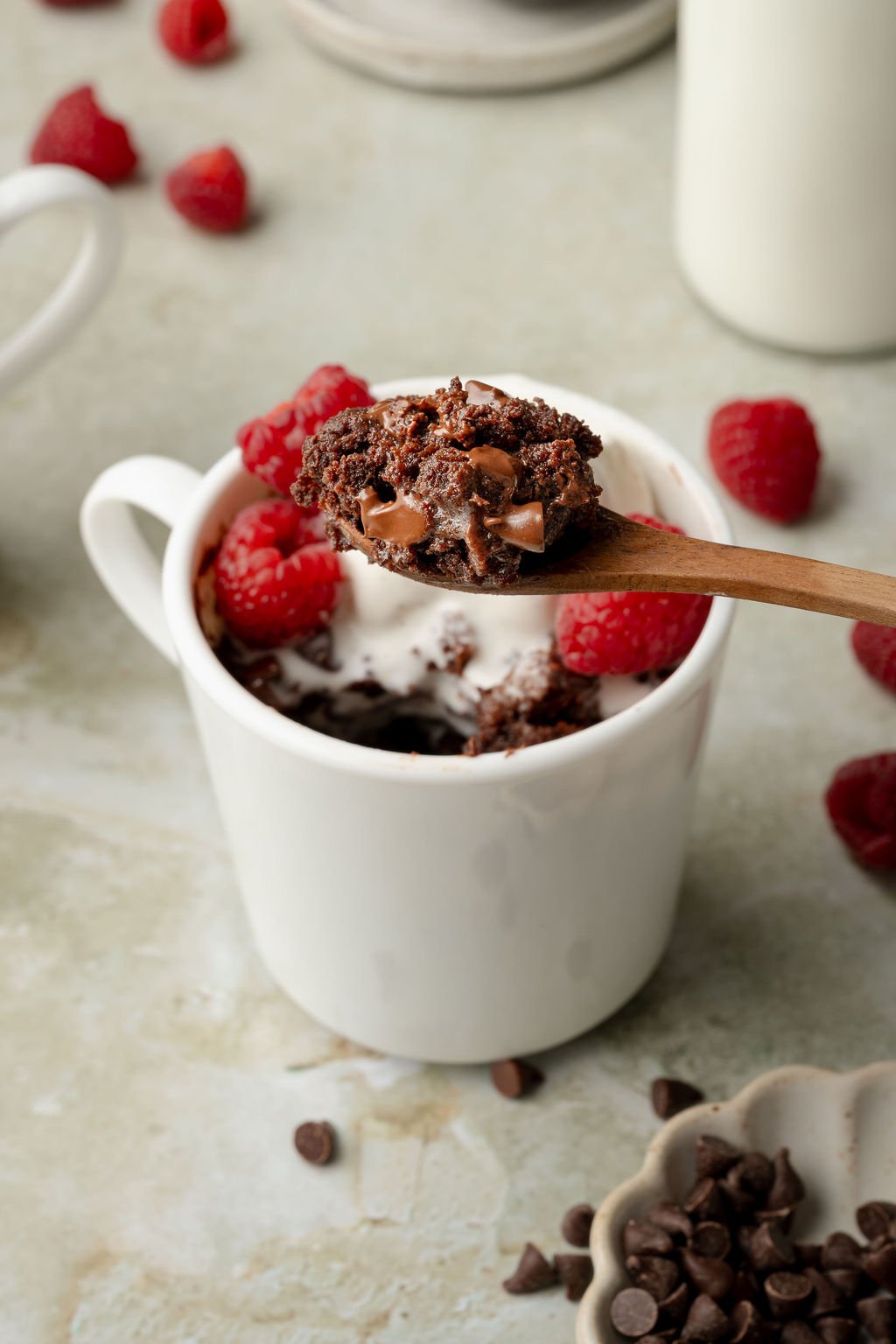 A close-up of a wooden spoon holding a gooey bite of chocolate mug cake, topped with melted chocolate chips. The mug is filled with chocolate cake, fresh raspberries, and a drizzle of cream.