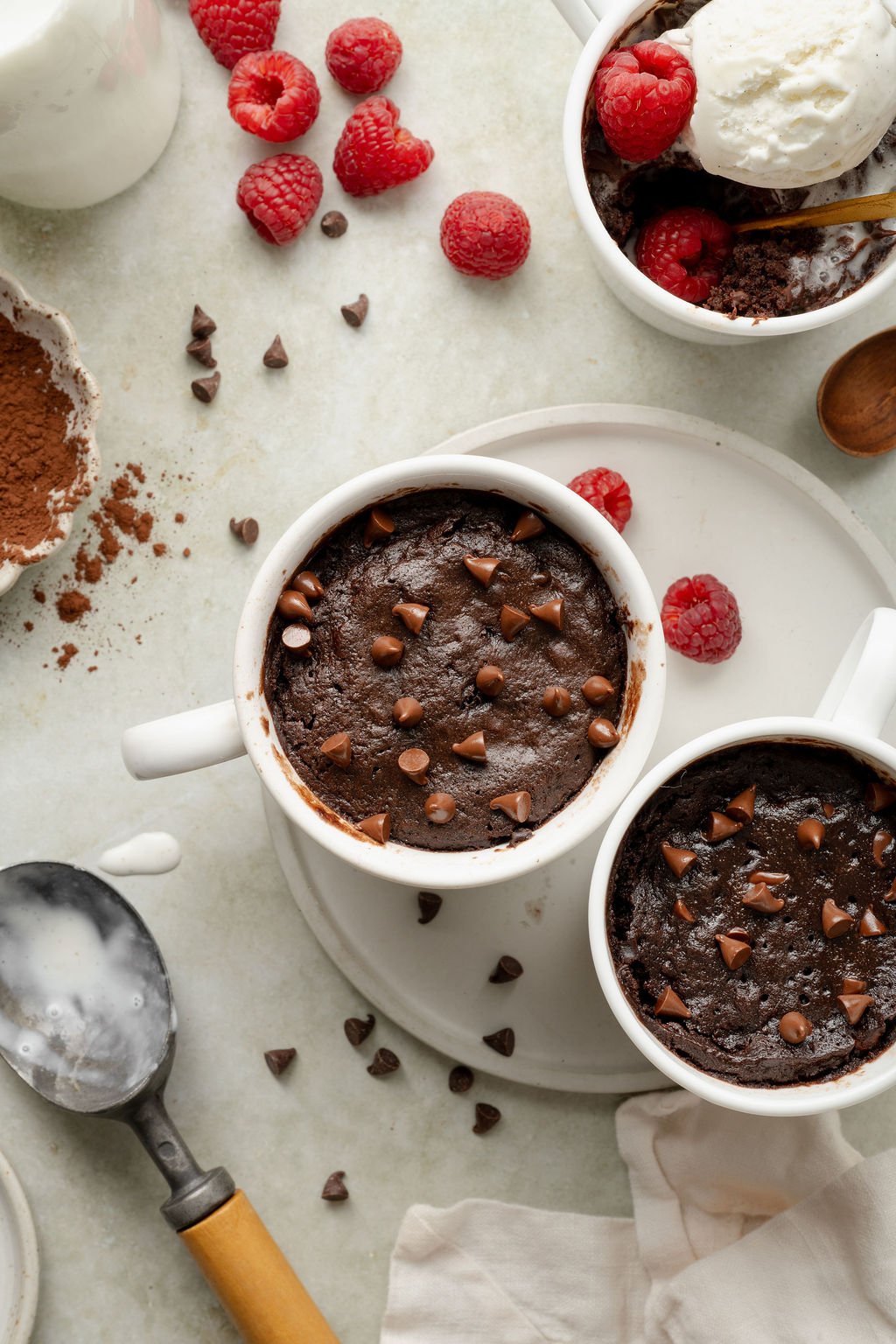 Overhead shot of two chocolate mug cakes in white mugs, topped with chocolate chips. Fresh raspberries, cocoa powder, and an ice cream scoop are scattered around on the tabletop.