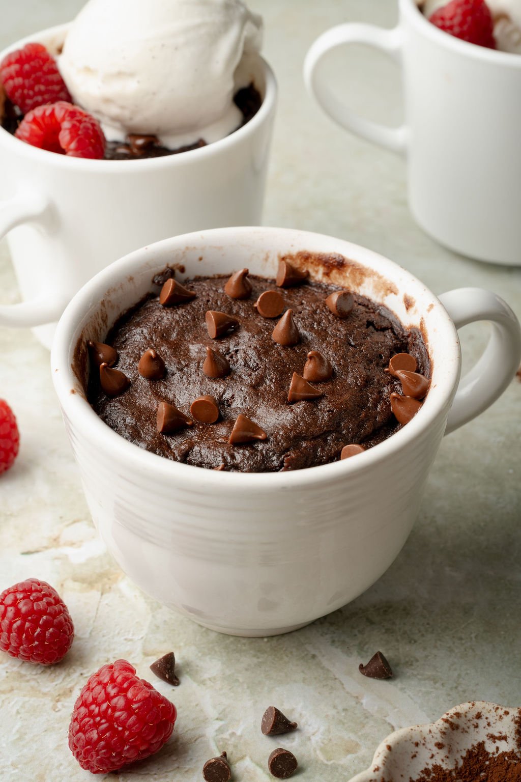 Side view of a freshly baked chocolate mug cake in a white mug, topped with chocolate chips. Raspberries and chocolate chips are scattered around for garnish.