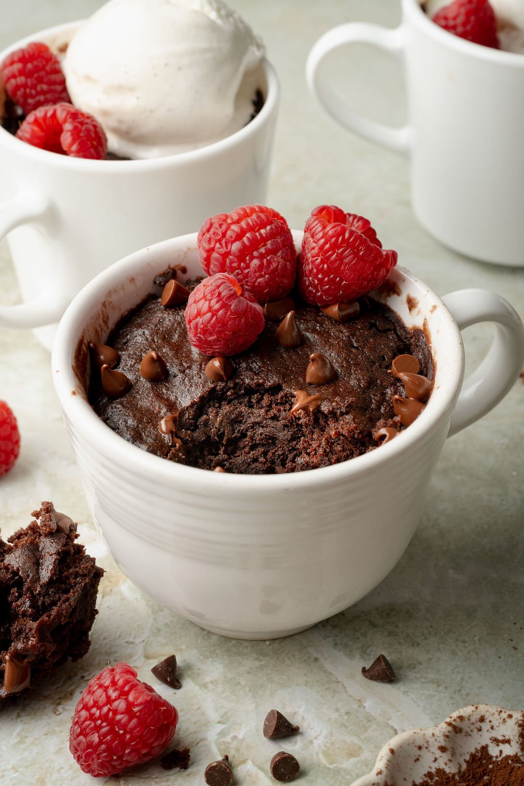 Chocolate mug cake in a white mug with a spoonful taken out, topped with fresh raspberries and melty chocolate chips. A second mug with ice cream is in the background.