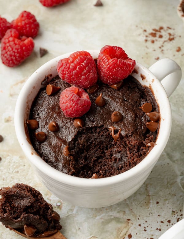 Close-up of a rich chocolate mug cake topped with raspberries and chocolate chips, with a bite missing and a spoonful of cake resting nearby.