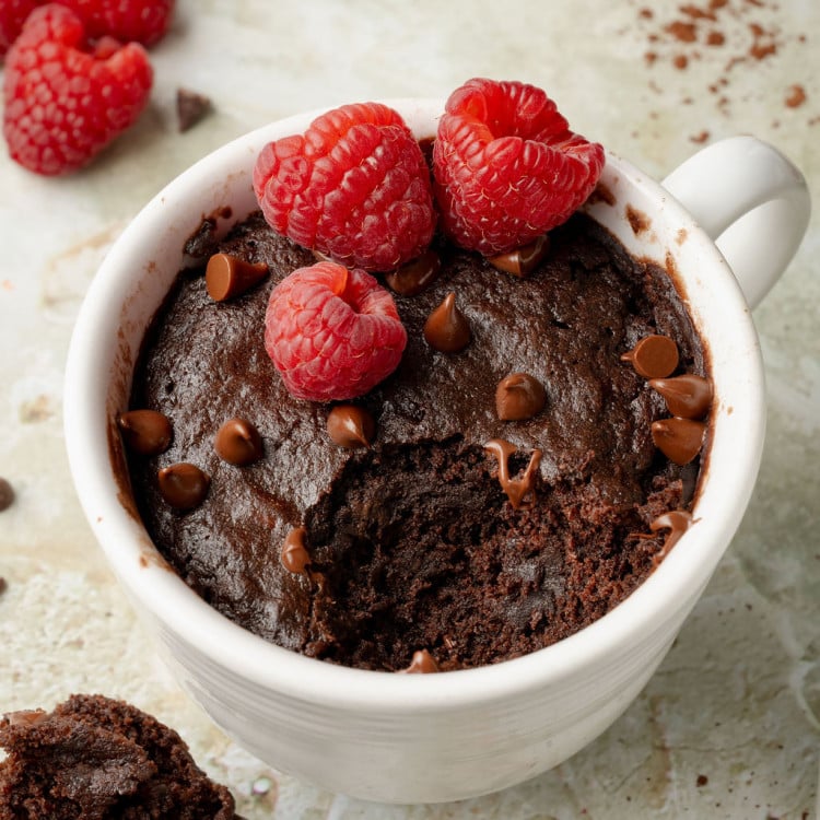 Close-up of a rich chocolate mug cake topped with raspberries and chocolate chips, with a bite missing and a spoonful of cake resting nearby.