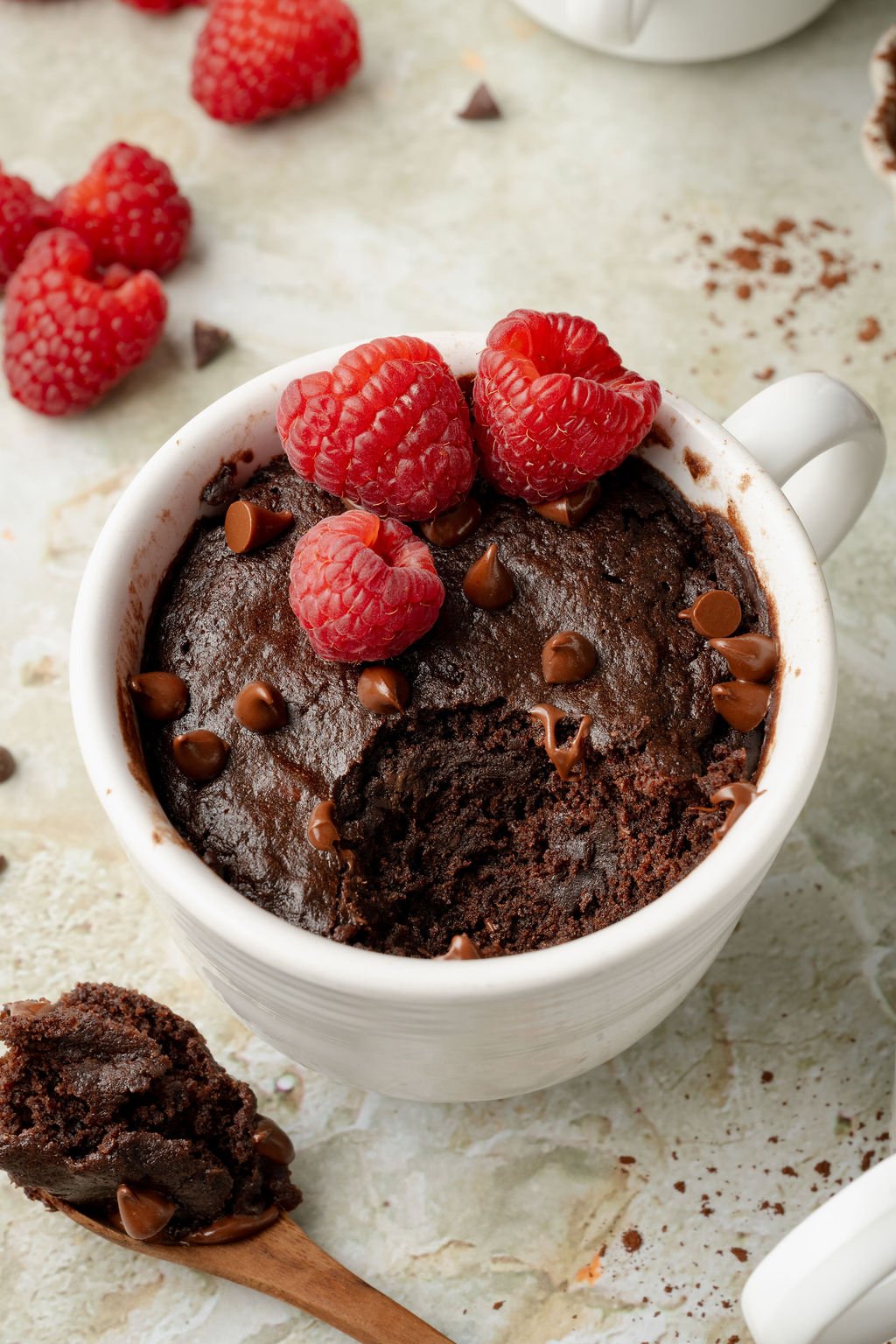 Close-up of a rich chocolate mug cake topped with raspberries and chocolate chips, with a bite missing and a spoonful of cake resting nearby.