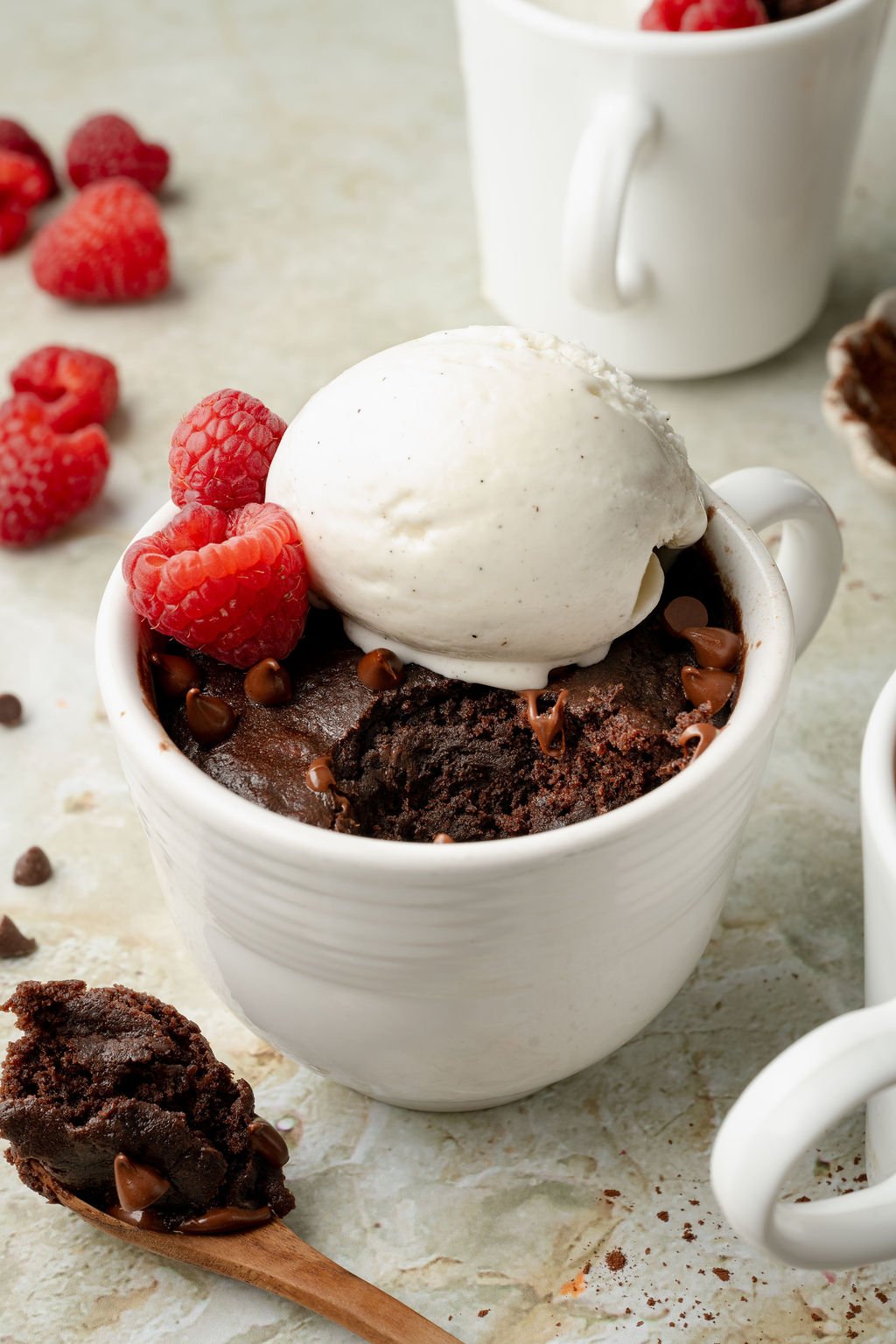 Chocolate cake in a white mug topped with vanilla ice cream and fresh raspberries. A spoonful of moist chocolate cake lies beside the mug on a marble surface.