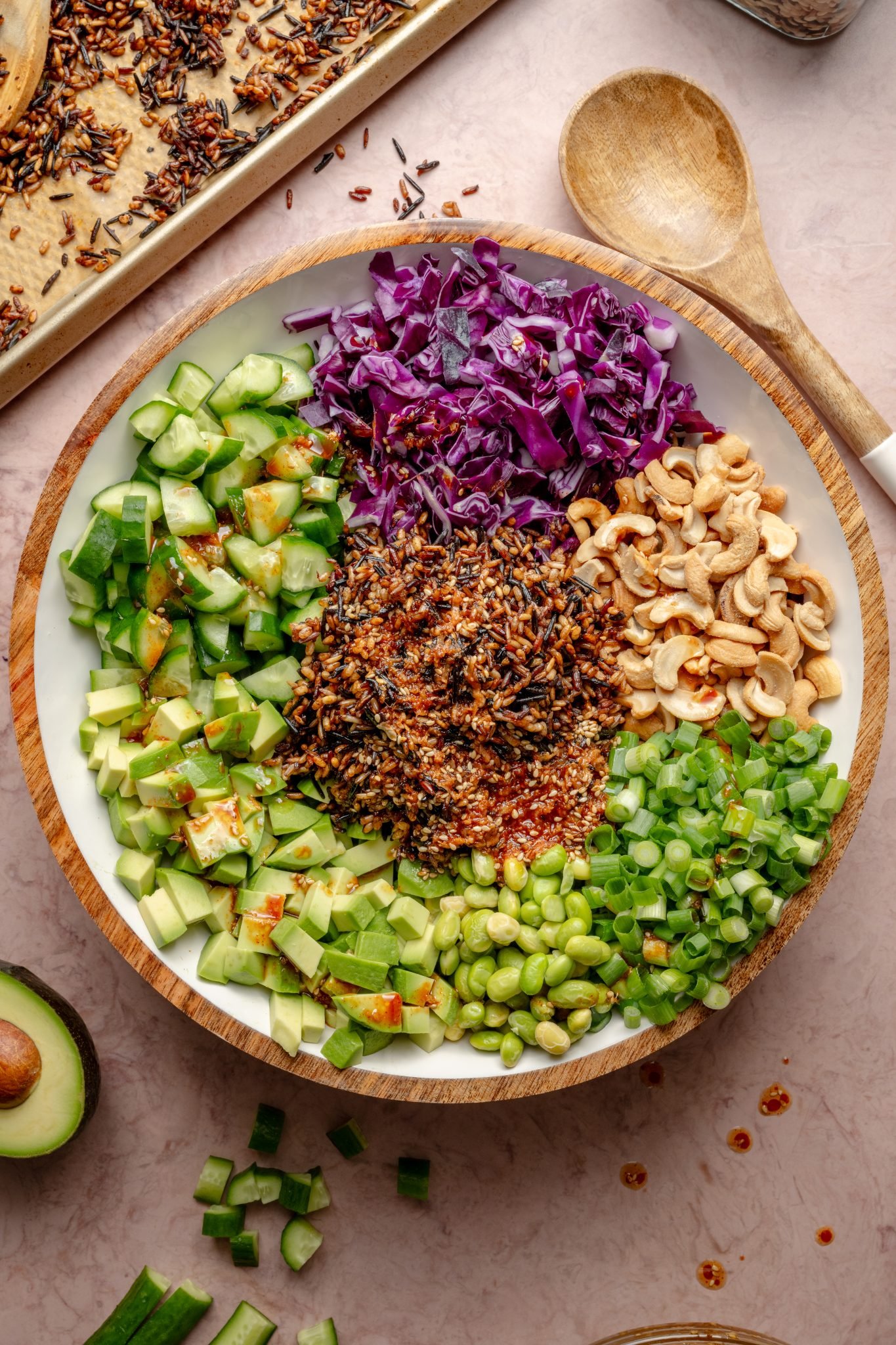 Overhead view of a large wooden bowl filled with colorful layers of chopped avocado, cucumber, purple cabbage, green onions, cashews, edamame, and crispy wild rice in the center, with a baking tray of toasted rice nearby.