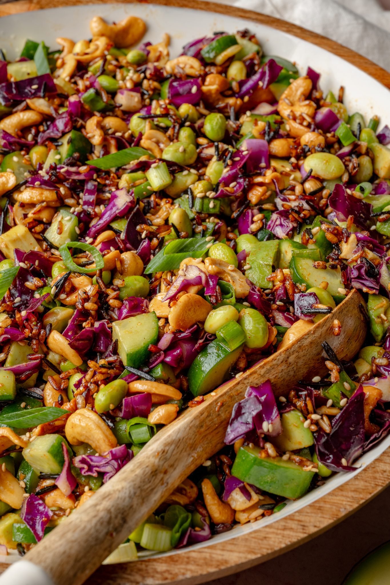 Close-up of the crispy rice salad showing a mix of textures and colors—crunchy cashews, chopped cucumbers, bright purple cabbage, and scattered sesame seeds with a wooden spoon in the bowl.