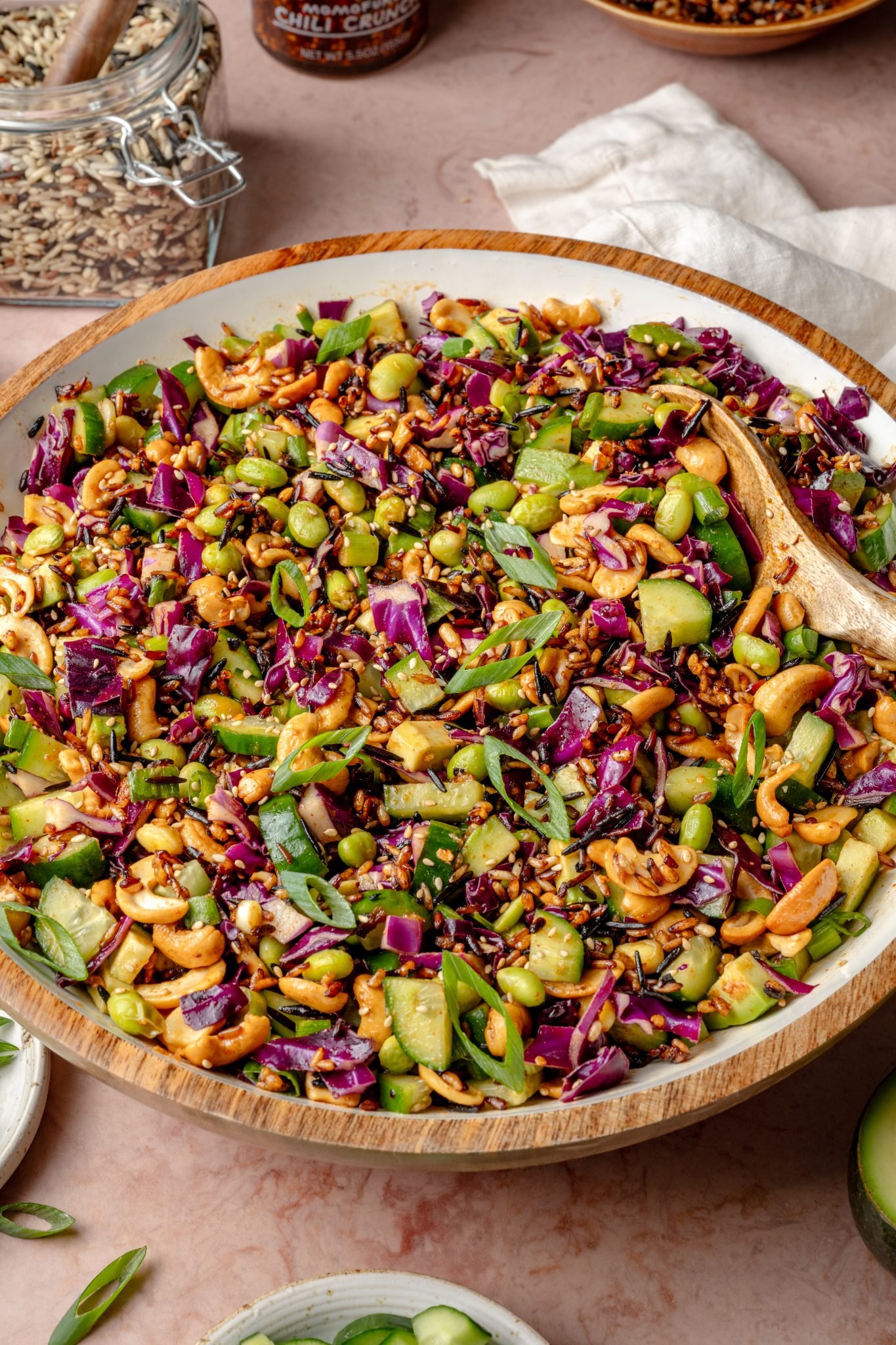 Large wooden salad bowl filled with tossed crispy rice salad, featuring vibrant purple cabbage, green onions, cucumbers, edamame, cashews, and sesame seeds, surrounded by dressing and ingredient jars.