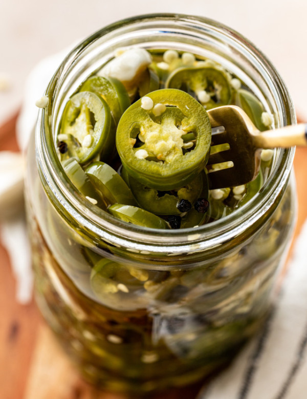 Pickled jalapenos in glass mason jar with gold fork lifting jalapeno out of jar.