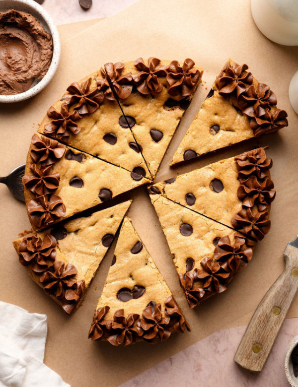 A sliced chocolate chip cookie cake decorated with piped chocolate frosting along the edges, with a cake server lifting one slice slightly away from the whole.