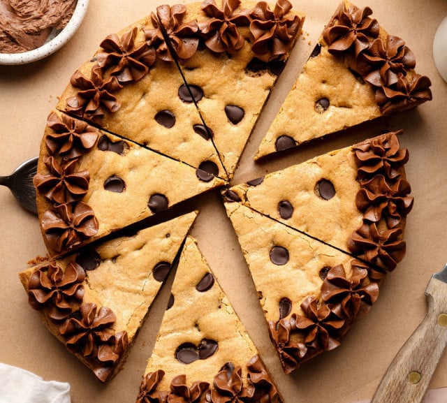 A sliced chocolate chip cookie cake decorated with piped chocolate frosting along the edges, with a cake server lifting one slice slightly away from the whole.